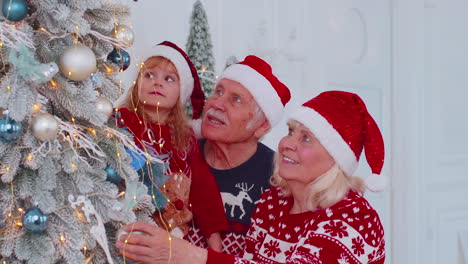 Toddler-girl-with-senior-couple-grandparents-decorating-artificial-Christmas-pine-tree-at-home-room