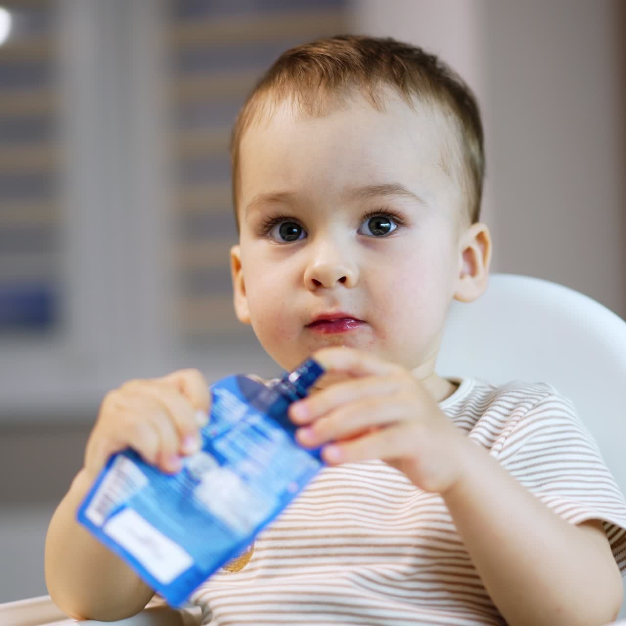 Lovely Caucasian toddler holds a pack of fruit puree in his hands. Baby tries to get some food from the pack with his finger. Close up. Blurred backdrop