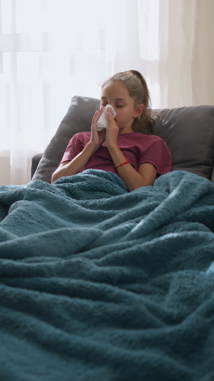 Female child in pink lying on couch suffering from flu, sneezing into tissue, with medication and glass cup on table, wrapped in blanket, looking unwell and tired