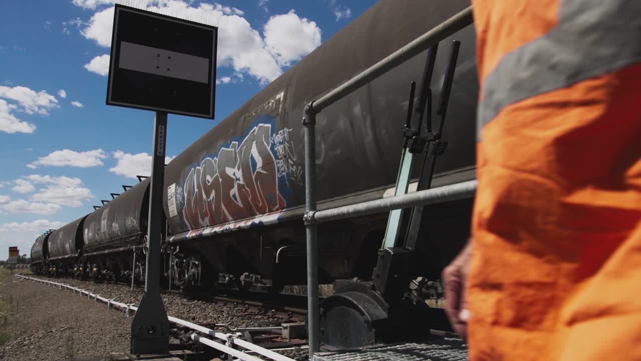 A freight train covered in graffiti passes over a manual railway turnout on a sunny day. A railway worker in high-visibility passes.