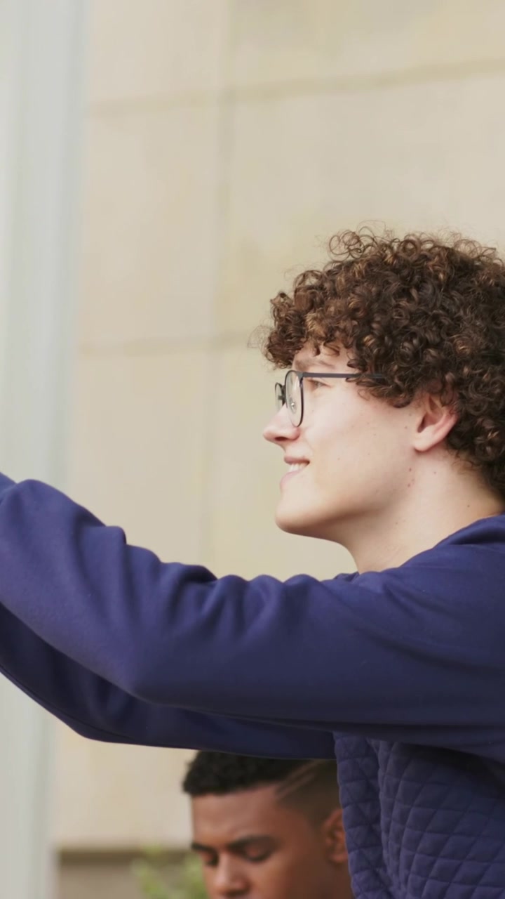 Video verticale: uomo che parla al telefono vicino al muro, bagaglio a mano accanto, compagno che entra e si fa un selfie