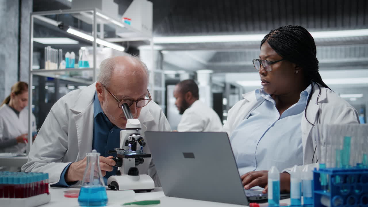 Pathologist examining biopsy slide while coworker documents findings on laptop