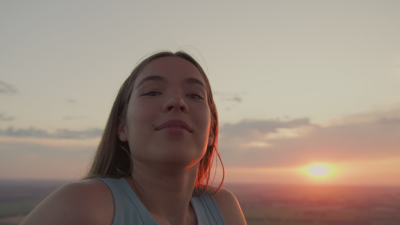 elegant young woman tilts head upward to watch metal burner flame flicker inside hot air balloon basket under warm sunset glow over sprawling fields and drifting clouds