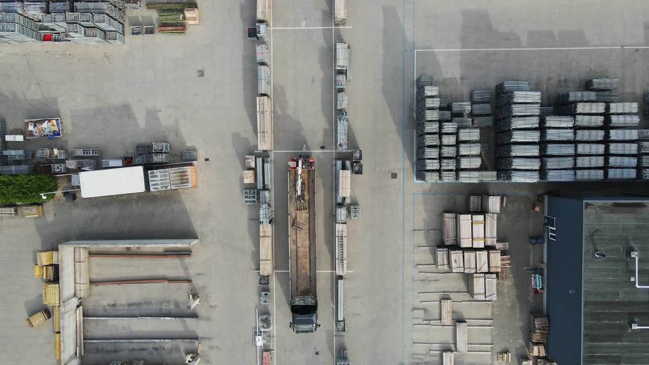 Top down aerial of truck with loading crane driving over an industrial storage yard filled with construction materials