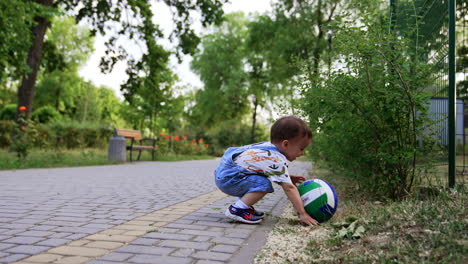 toddler boy in jeans romper wants to lift the ball from the ground. baby misses a little, nearly falls and looks at his palm.