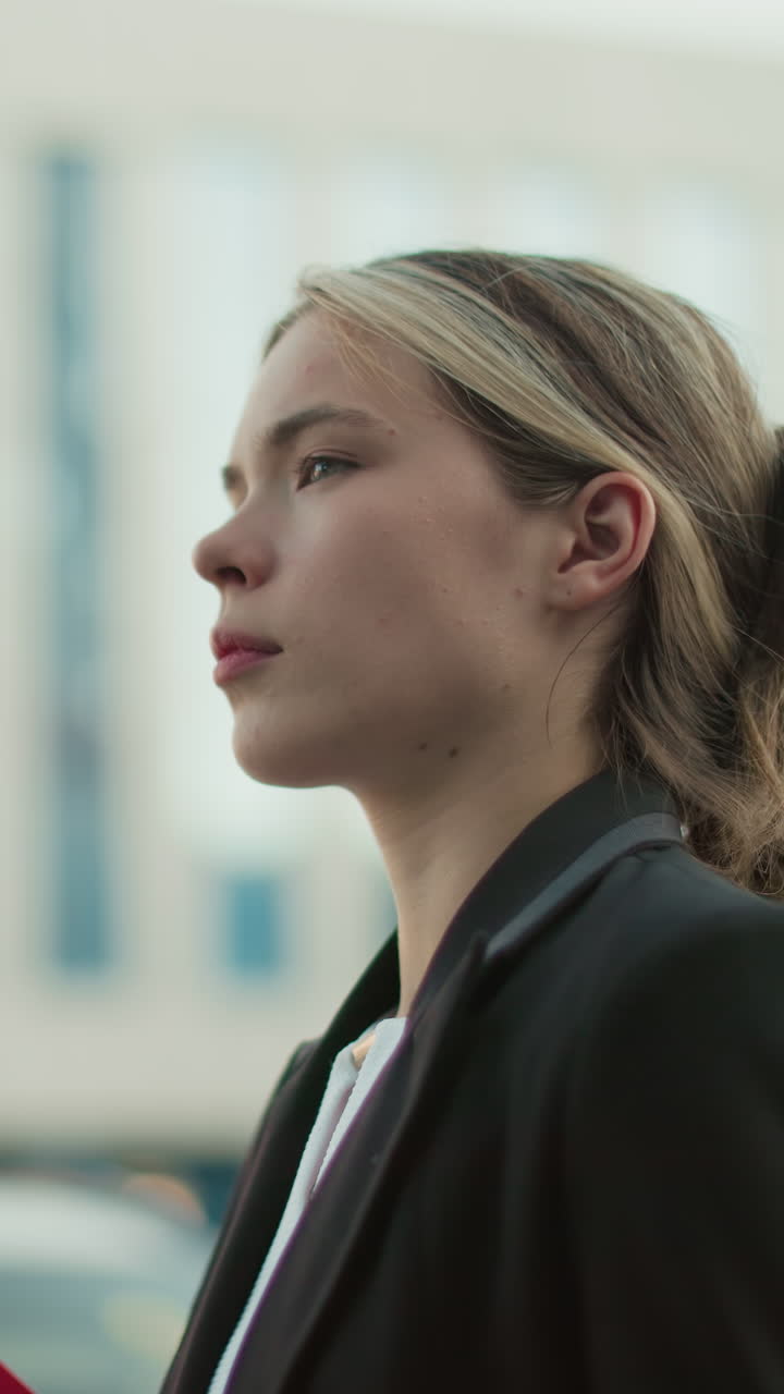 Elegante receptionist che cammina con una cartellina rossa in mano, guarda verso destra con lo sfondo sfocato di un alto edificio moderno e auto parcheggiate in città durante una giornata lavorativa luminosa in un ambiente urbano professionale
