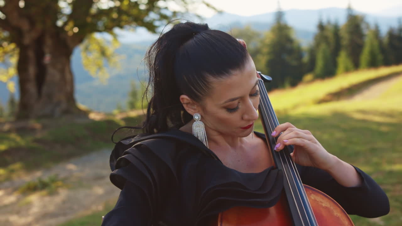 Young adult woman performing her cello play at the nature. Female music player closes her eyes enjoying music. Beautiful sunny day nature backdrop.