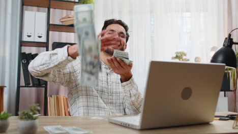 Happy rich indian office businessman working on laptop wasting throwing money dollar cash earnings