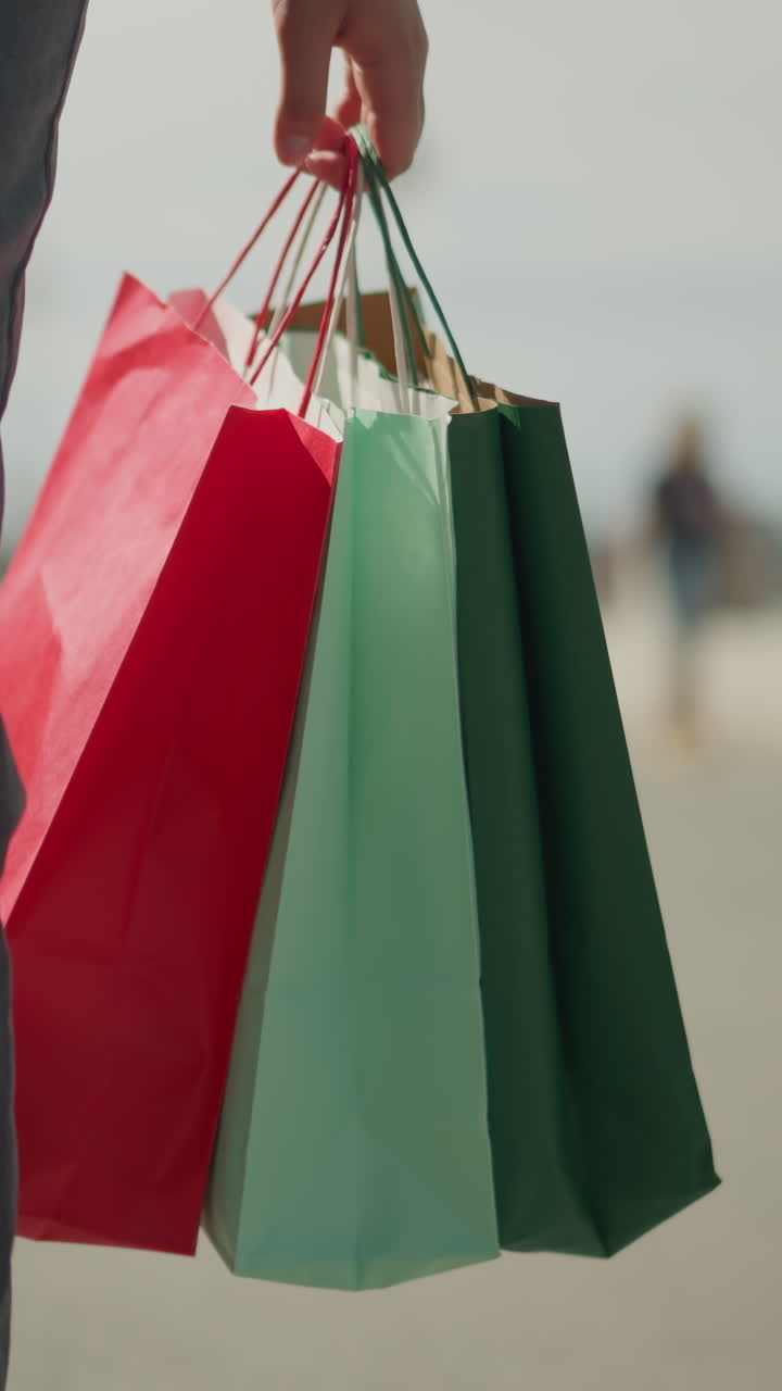 vista de la pierna de una persona caminando por una calle sosteniendo bolsas de compras coloridas, con una mano visible llevando un bolso de mano, el fondo borroso presenta a otros transeúntes y un aire libre ligeramente urbano