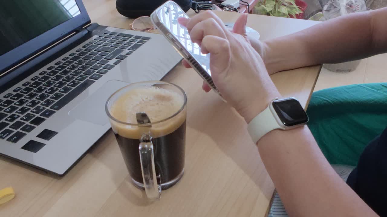 Close-up on hands using smartphone, with laptop, coffee, and smartwatch.