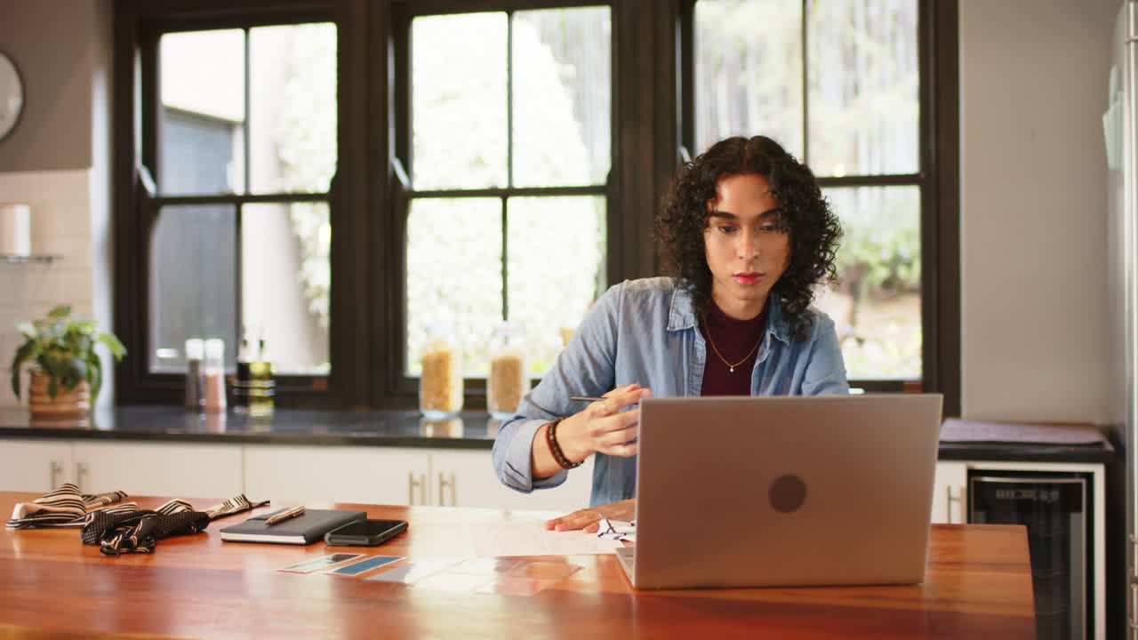 Non-binary person scanning laptop, reaching for pen, jotting notes for work at kitchen