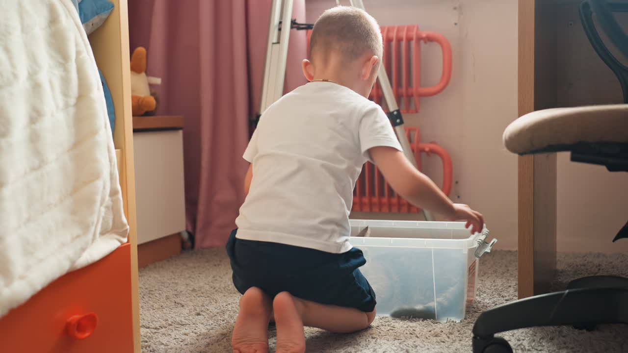 White son searching toy container on carpet, kneeling and leaning into clear plastic box, soft morning light through curtain, candid home moment of playful curiosity and childhood discovery