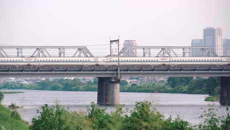 shinkansen bullet train travelling across the bridge of a calm tamagawa river with city landscape at the background during daytime in tokyo, japan