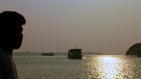 Un-Hombre-Indio-Se-Enfrenta-A-La-Cámara-A-Orillas-Del-Lago,-Al-Atardecer,-Casas-Flotantes-Al-Fondo,-Silueta