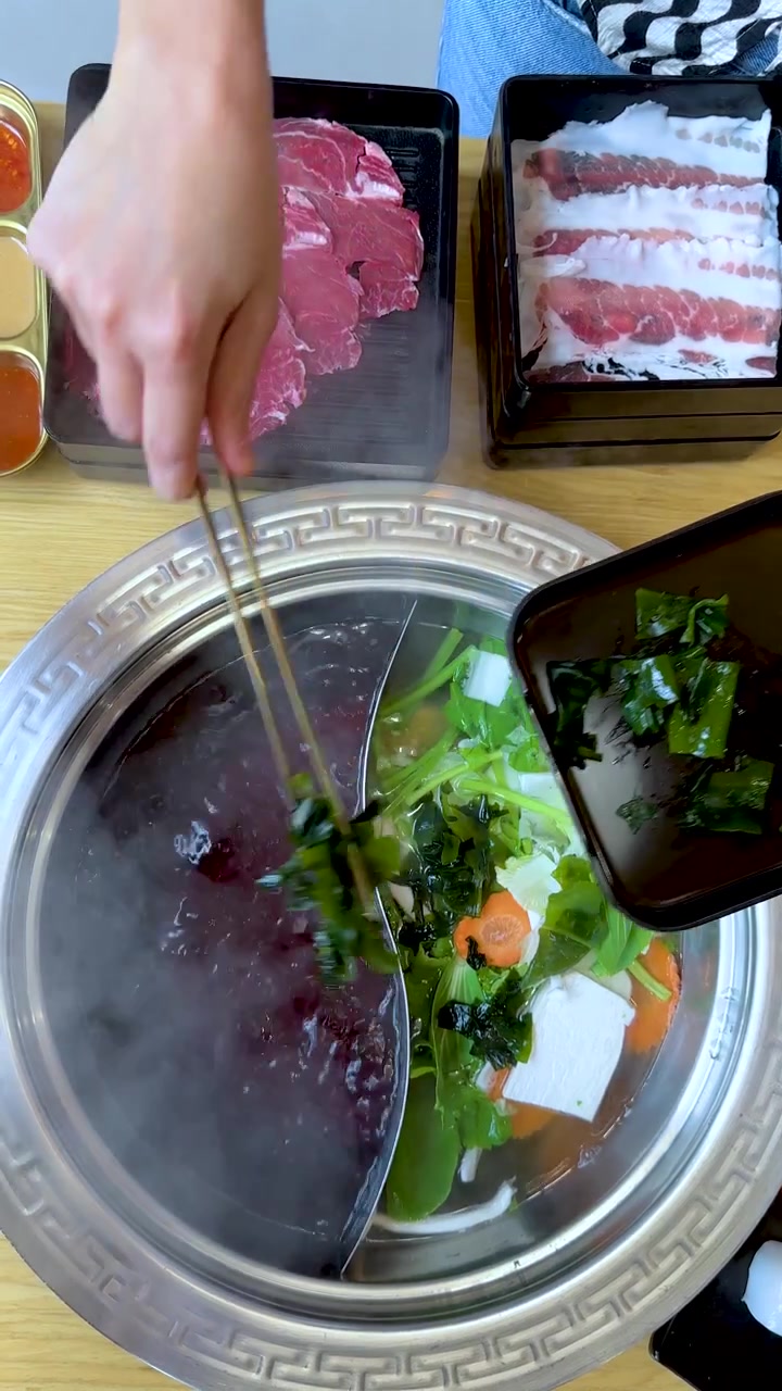 スライス牛肉、豆腐、青菜が入った湯気の立つ二色鍋の火鍋に、手が野菜を加える。