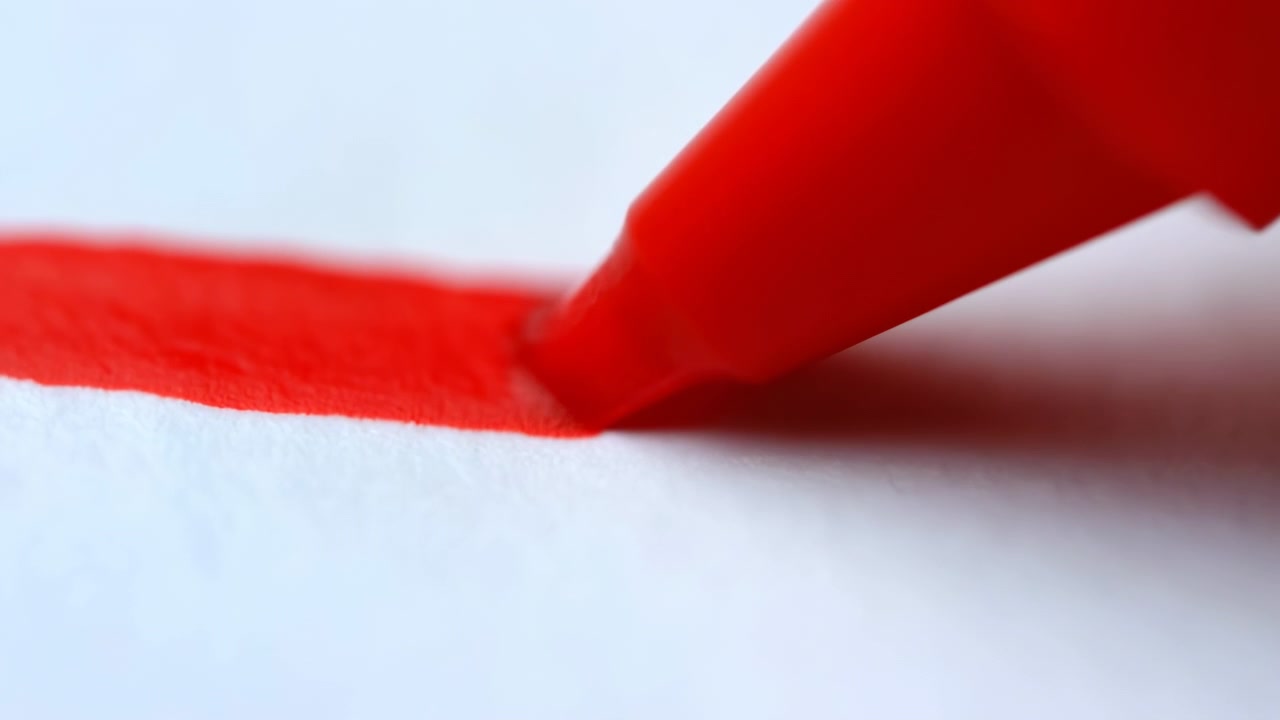 Primo piano: punta di pennarello rosso che disegna una linea marcata, saturazione dell'inchiostro nitida su carta bianca testurizzata, punta angolata precisa.