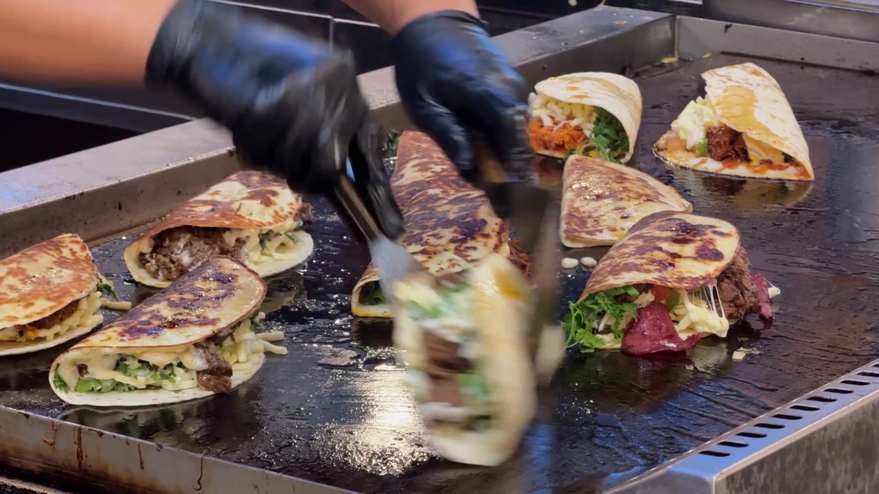 Filmagens em close-up de um vendedor de comida preparando vários tacos e wraps frescos numa chapa quente. Tortillas são recheadas com carne, queijo, vegetais, ervas e molhos durante a movimentada preparação de comida de rua.