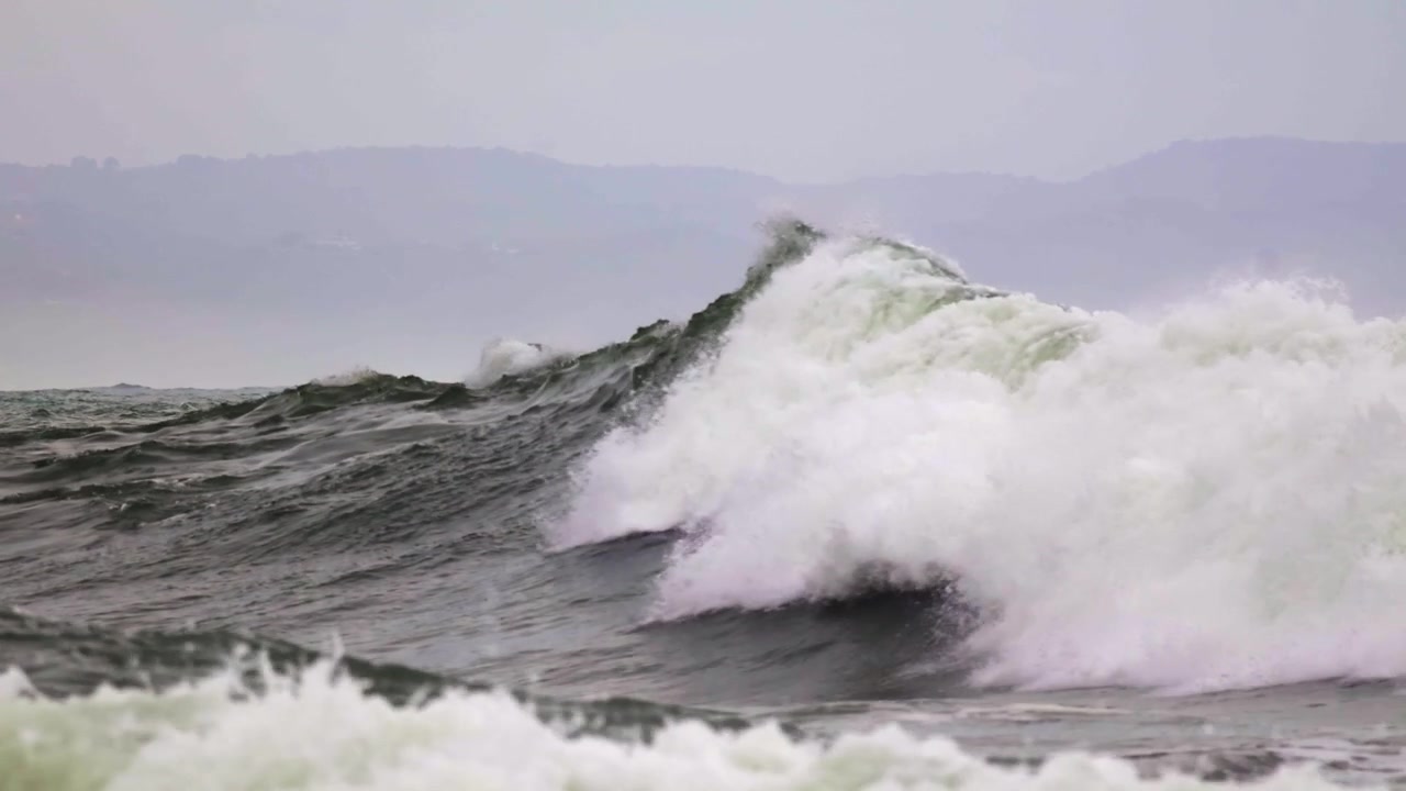 Large deep-sea swell building and crashing with turbulent white water across the frame. Fixed tripod telephoto shot. Misty, hazy coastal hills in the distant background under an overcast sky.