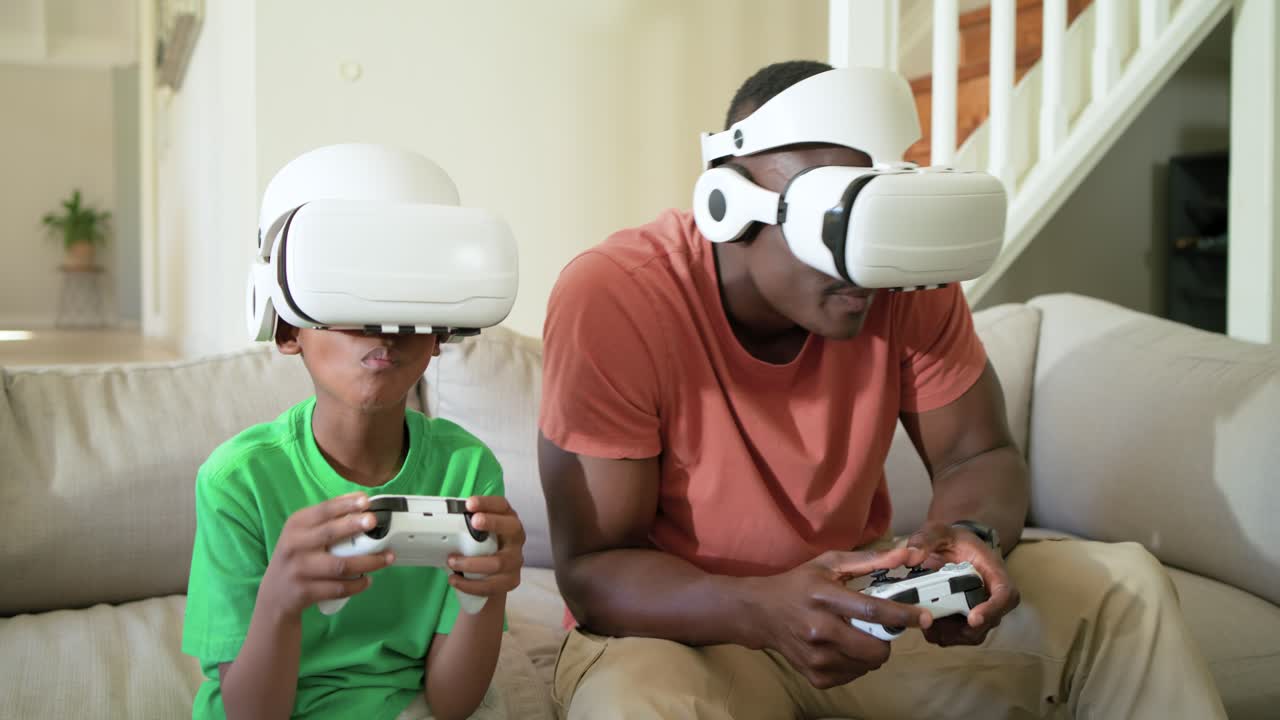 Padre e figlio afroamericani indossano visori VR e reagiscono a un gioco VR a casa, impugnando i gamepad.