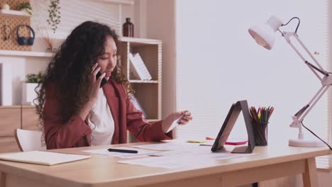 la mujer diseñadora asiática usa su teléfono inteligente para comunicarse, trabajar en proyectos y papeles, y trabajar desde casa en un escritorio con una computadora portátil.