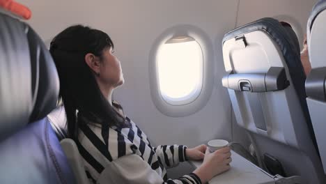 Woman-sitting-inside-airplane-and-looking-at-window-and-drinks-coffee.