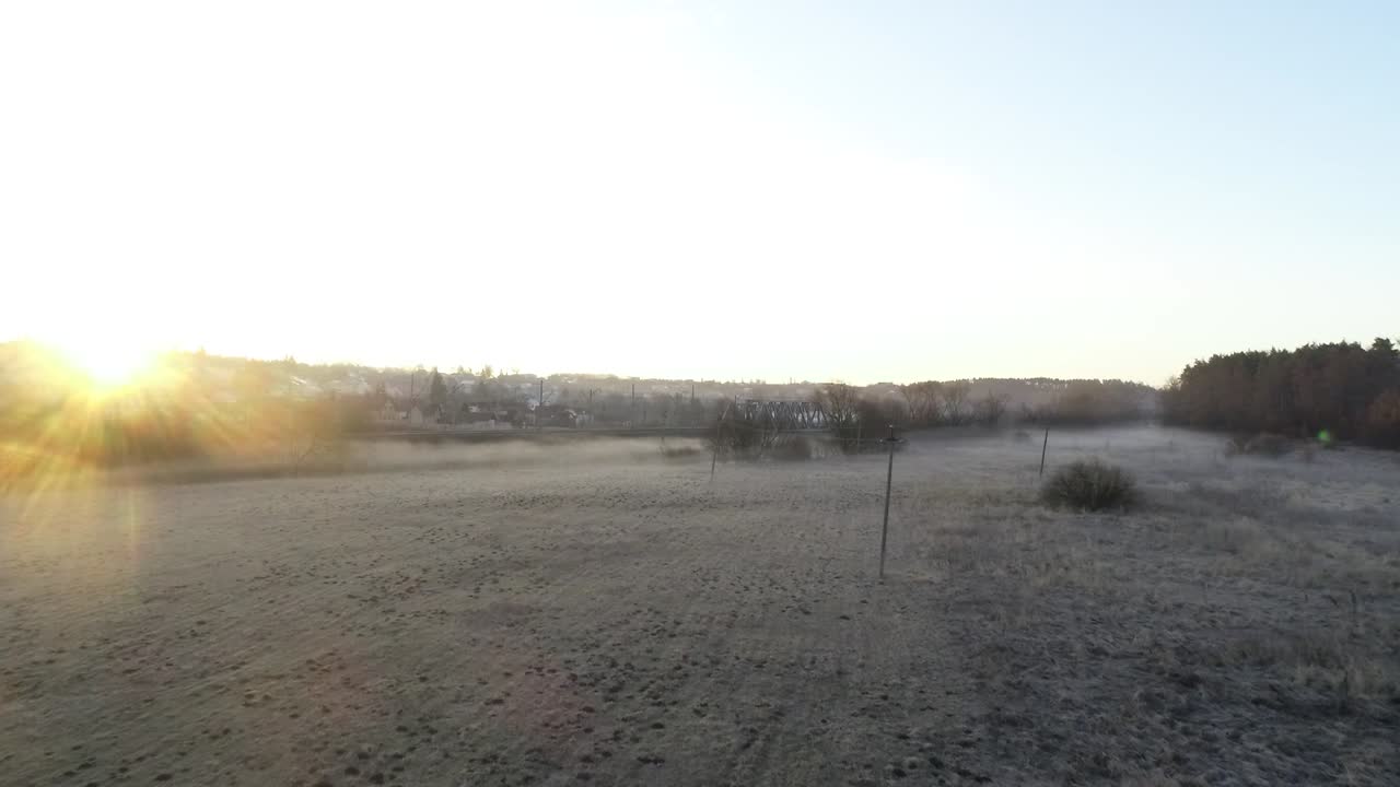 Early morning, the sun is rising over the village where there is a railway and near an empty field covered with fog and frost lies on the grass. The weather is fine and the sky is blue!