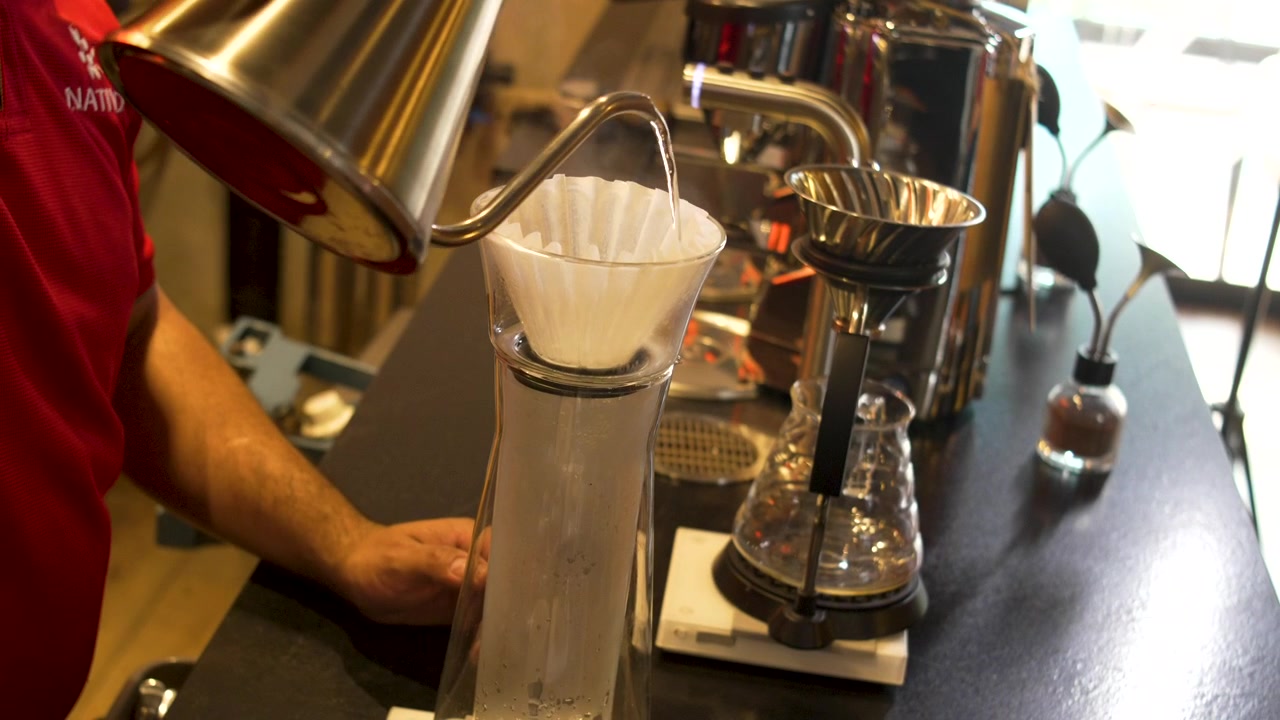 Preparazione manuale del caffè con metodo pour over: bollitore in acciaio che gocciola acqua su filtro di carta in una caffetteria specializzata.