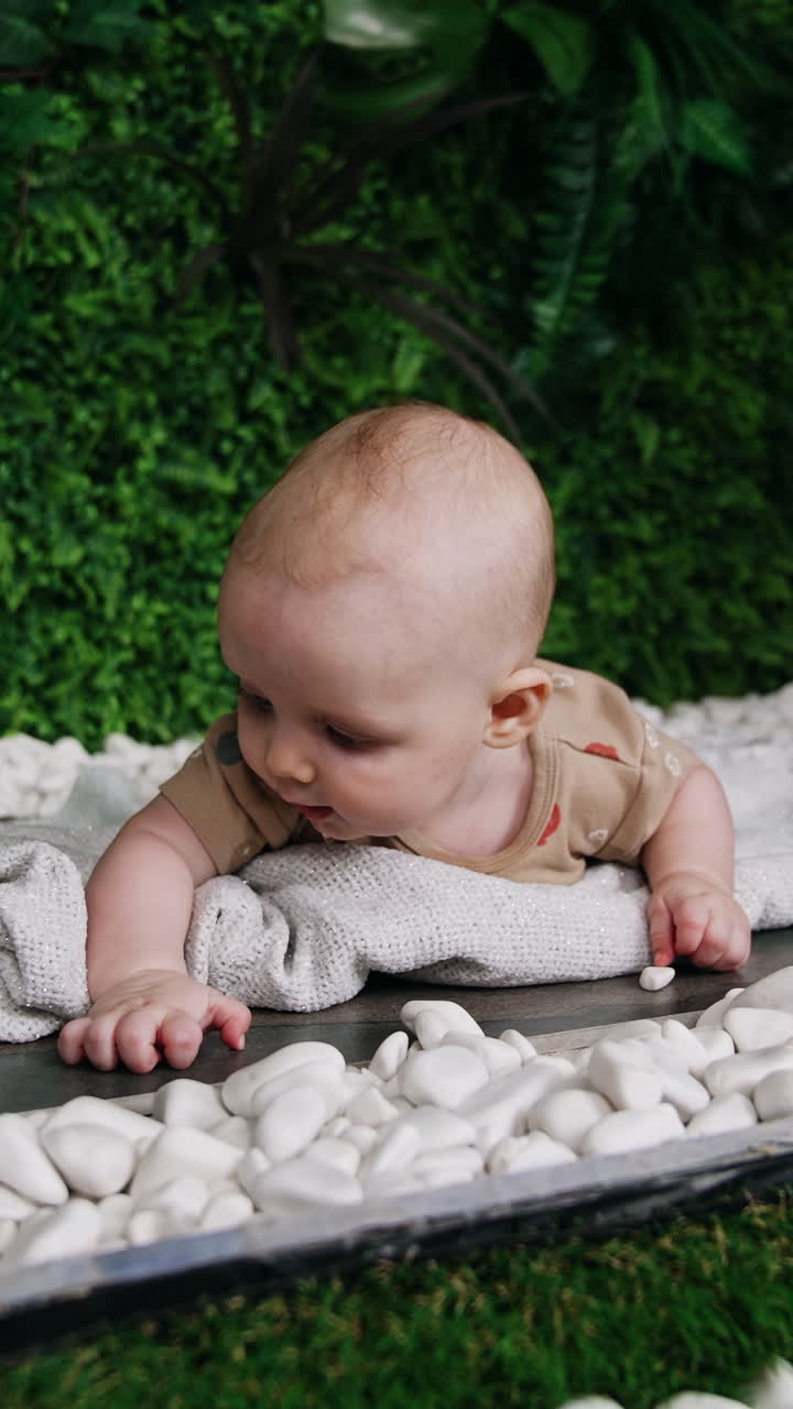 Caucasian baby boy lies on the white plaid. Cute kid is interested with white pebbles around him. Artificial greenery at backdrop. Vertical video