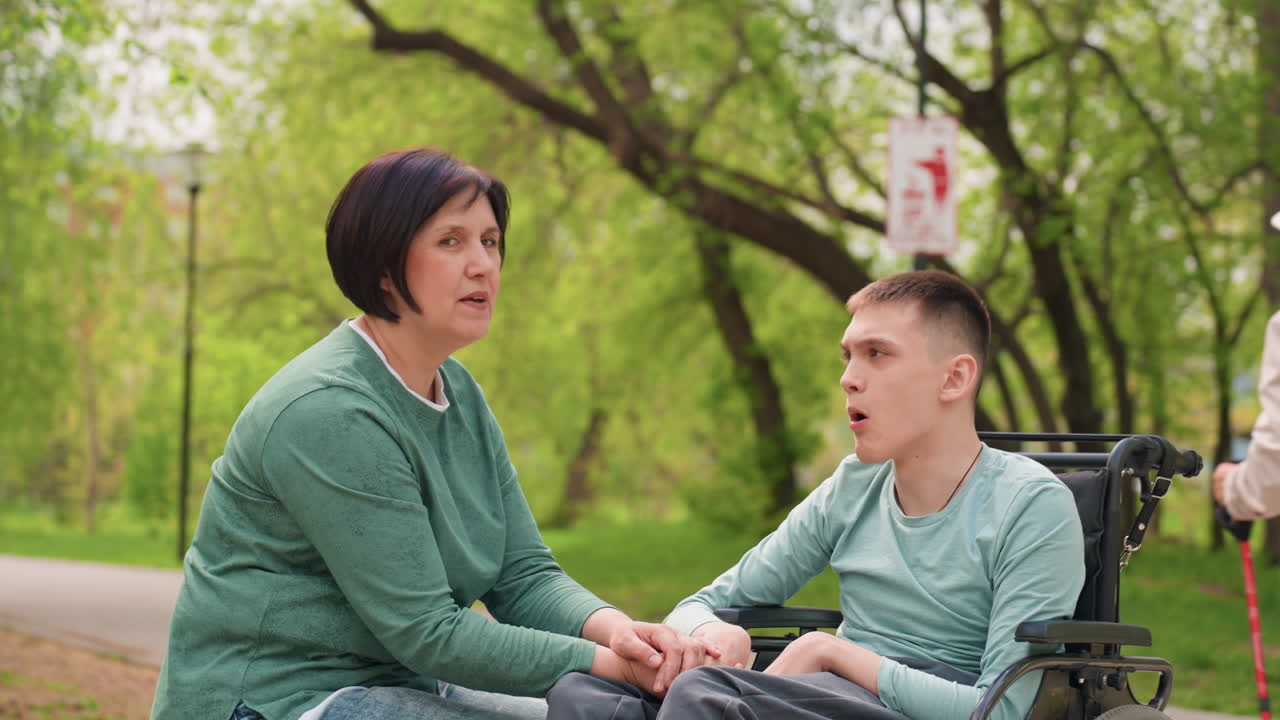 Mother And Son Near Wheelchair Sharing Heartfelt Conversation On Bench, Tender Holding And Expressive Communication, Green Trees And Soft Spring Light Create Warm Empathetic Atmosphere