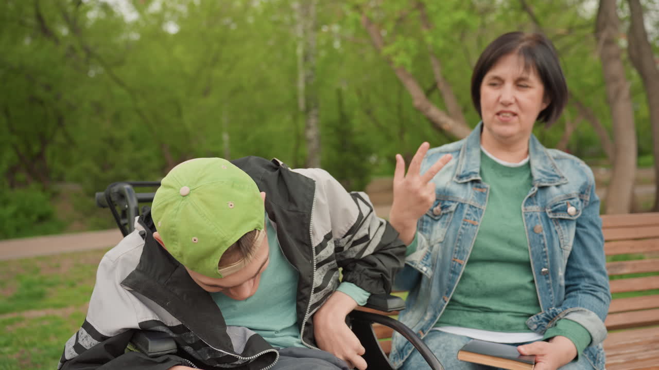 Gentle Assistance By Woman For Young Man, Compassionate Woman Helping Young Man Sitting On Park Bench, Caring Woman Guiding Young Man On Wheelchair In Relaxing Outdoor Park Setting