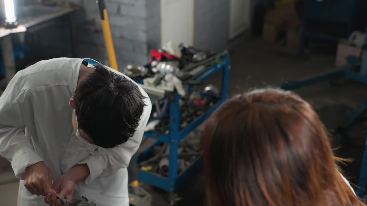 vista aérea de un estudiante en un taller automotriz que utiliza una pinza de vernier para medir un componente del motor mientras muestra el registro de medición a un colega