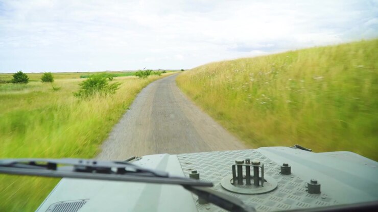 Una prospettiva avventurosa in prima persona alla guida di un robusto veicolo utilitario attraverso verdi campi agricoli. La strada sterrata e sconnessa davanti suscita una sensazione di pura eccitazione e libertà assoluta.