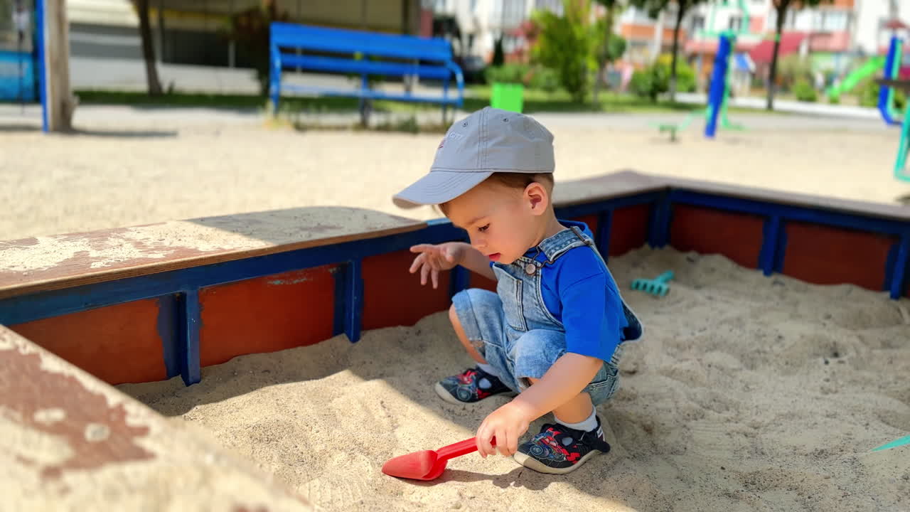 Little Caucasian toddler in jeans romper playing in sand box. Baby boy drops a shovel and takes sand with hands.