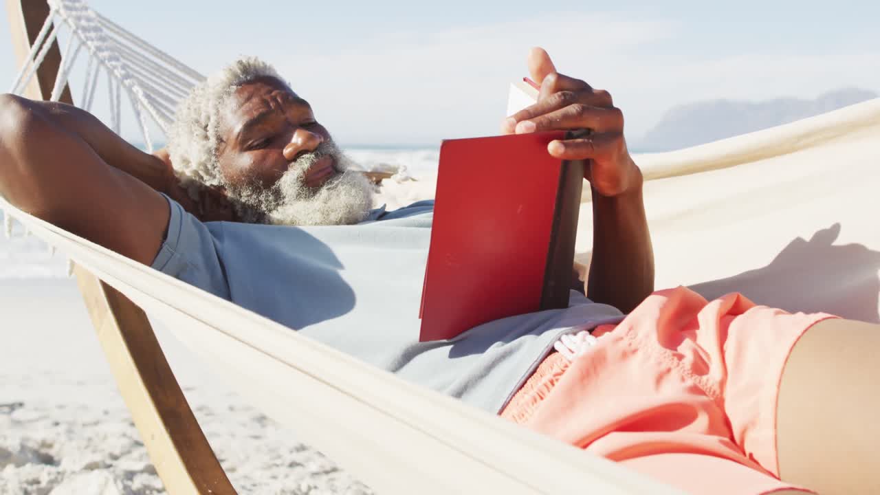 hombre afroamericano mayor feliz leyendo y acostado en una hamaca en una playa soleada