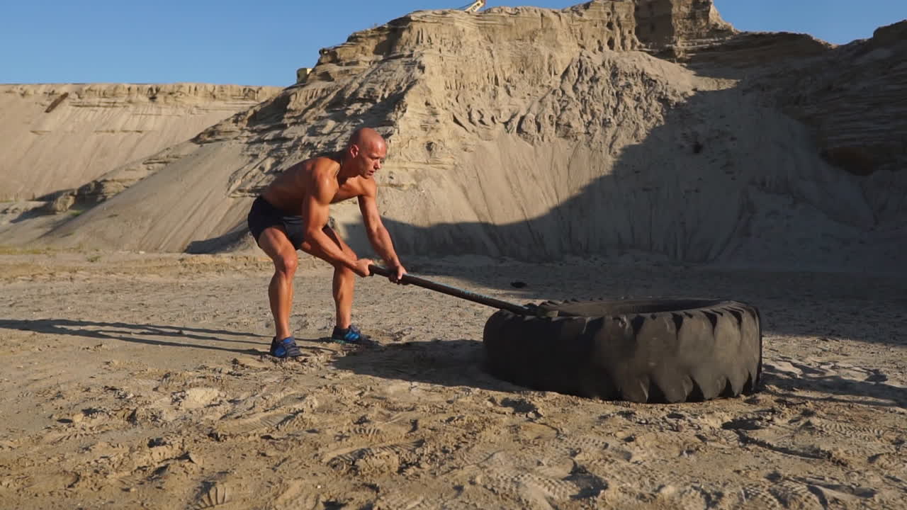 근육 운동선수 강인한 남자가 모래 산에서 커다란 바 ⁇ 에 망치를 때리며 서서히 일몰합니다. 바 ⁇ 의 먼지가 올라갑니다.