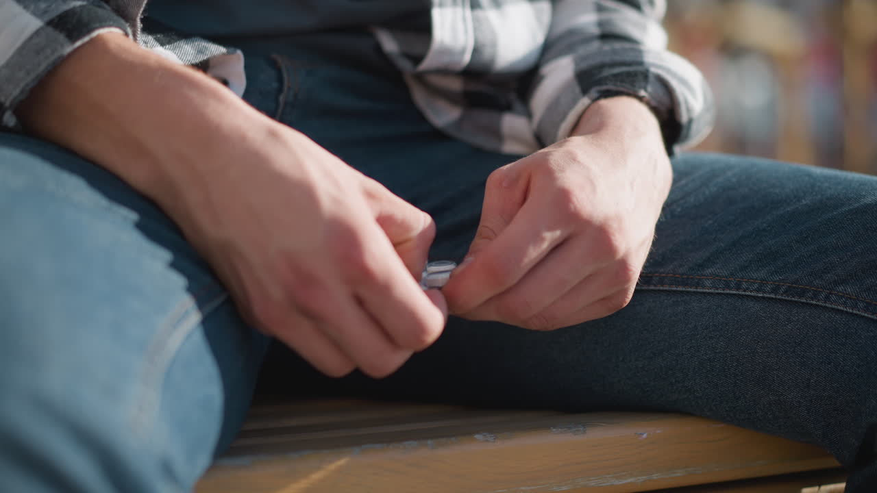 close up of hands opening medicine pack while seated on wooden bench wearing denim jeans and checkered shirt with bright outdoor lighting and soft blur background of park greenery