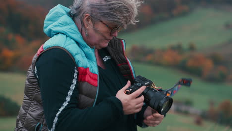 una mujer fotógrafa mirando a su cámara, sonriendo y tomando fotos, el viento jugando con su cabello gris en un paisaje de otoño con coloridas hojas de naranja