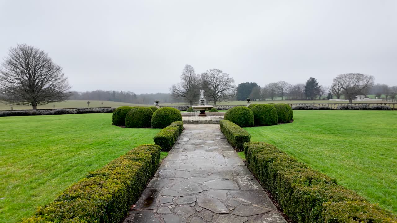 Ein formeller Gartenweg, der an einem trüben Tag zu einem Brunnen führt, mit einem grünen Rasen, geschnittenen Hecken und einem Steinweg in einer ruhigen Außenanlage für Landschaftsgestaltungskonzepte