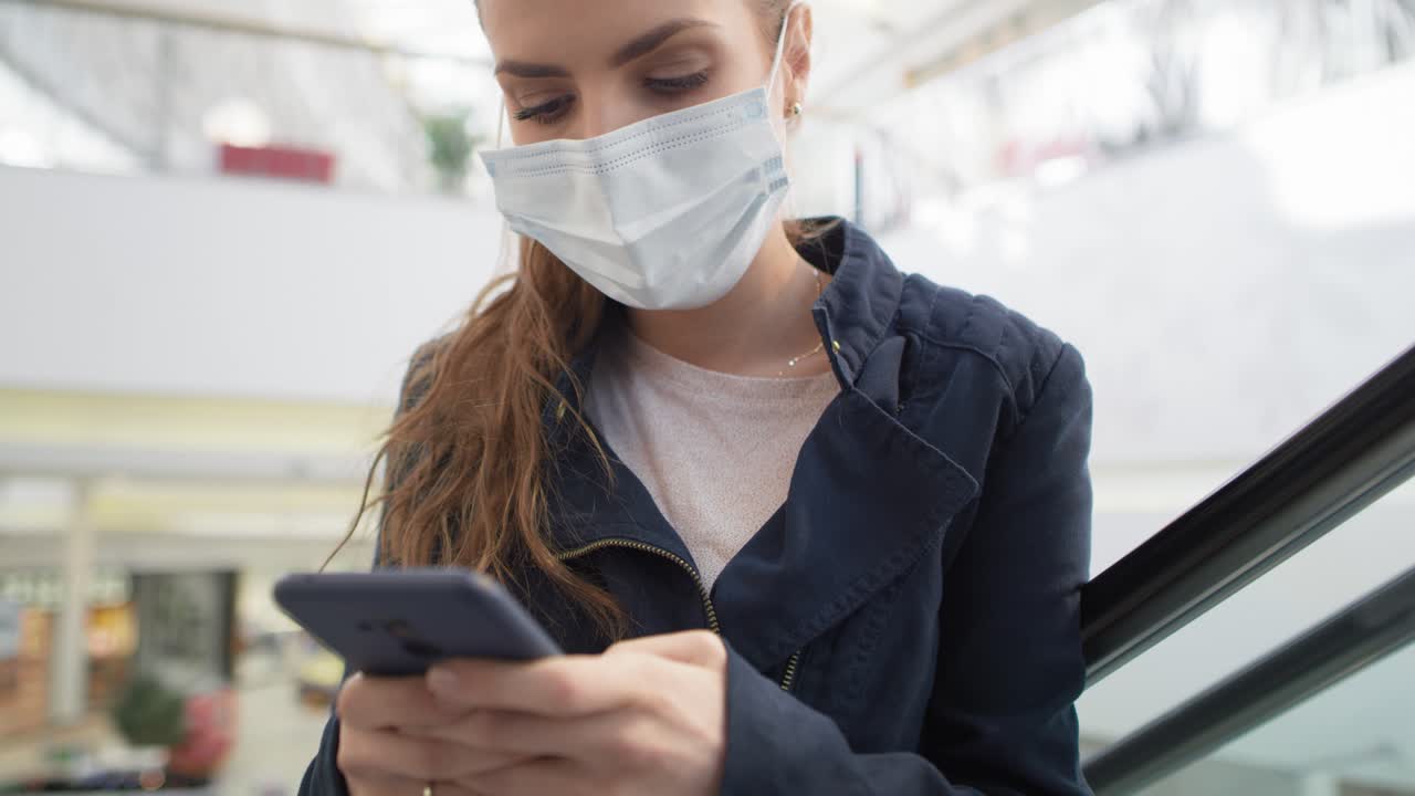 video de una mujer con máscara médica usando un teléfono inteligente en una escalera mecánica