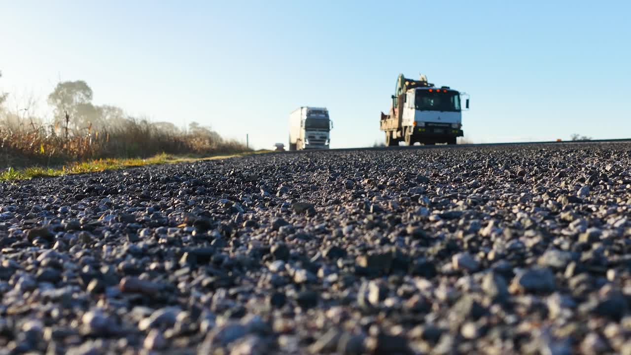 A truck and car approach on a gravel countryside road in early morning light, captured from a low ground-level perspective with a static camera