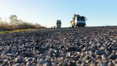 a truck and car approach on a gravel countryside road in early morning light, captured from a low ground-level perspective with a static camera