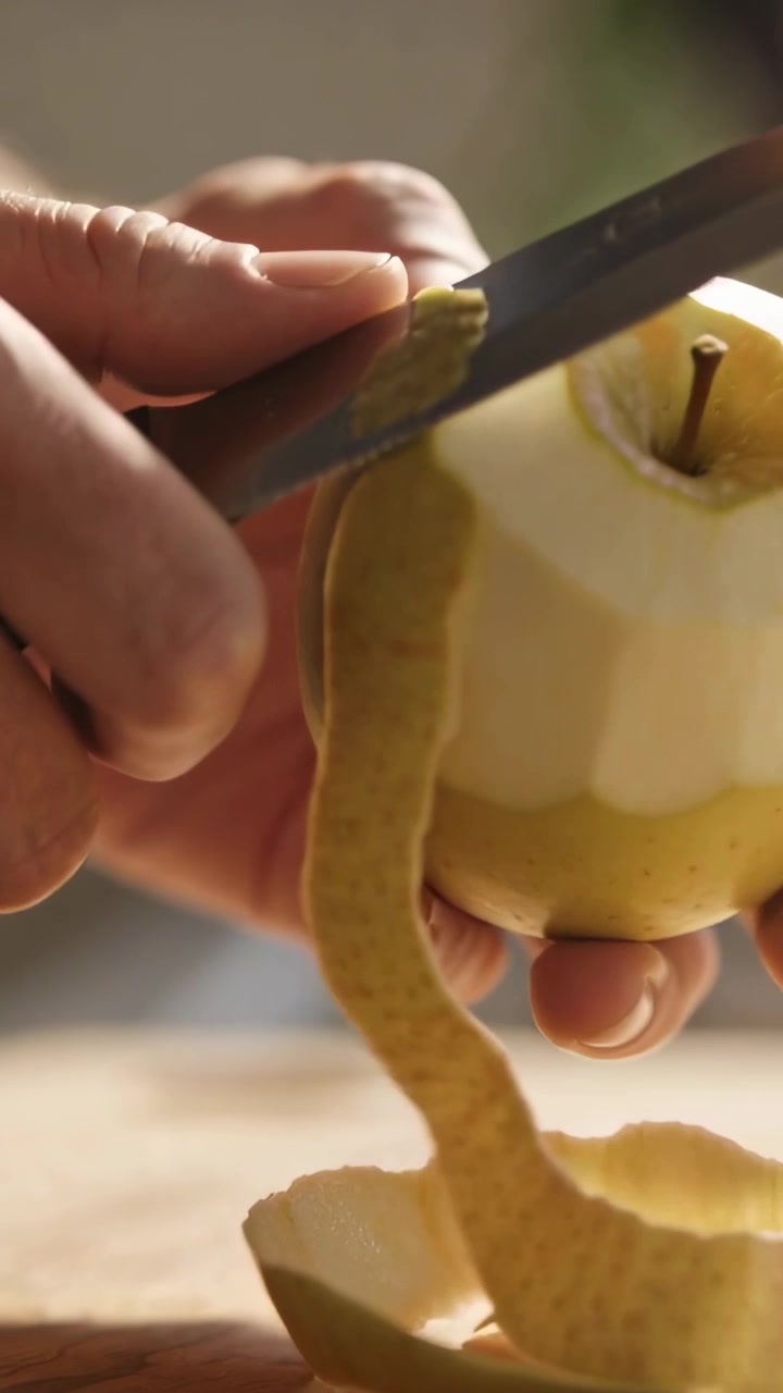 Video verticale: Mani che sbucciano una pera con un coltellino, affettando sotto il picciolo in cucina, preparando la frutta.