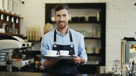 Porträtaufnahme-Des-Gutaussehenden-Barkeepers,-Der-Tassen-Kaffee-Hält-Und-Zur-Bar-Geht,-Während-Seine-Kollegin-Hinter-Der-Kaffeemaschine-Mehr-Zubereitet