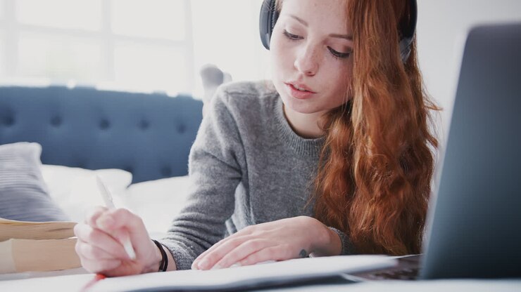 Kvindelig universitetsstuderende med hovedtelefoner arbejder på sengen i et delt hus med bærbar computer og mobiltelefon