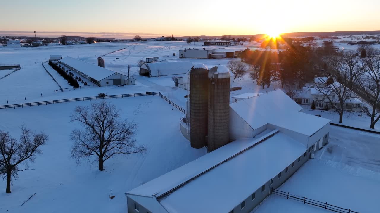 Snedækket gård med lader, hus og siloer under vinterens solnedgang i den gyldne time
