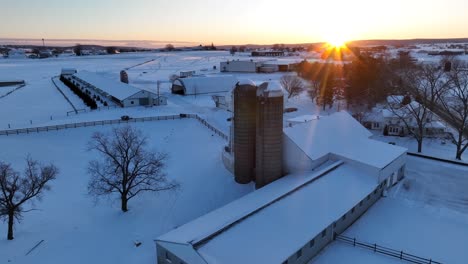 snow covered farm with barns, house, and silos during golden hour winter sunset