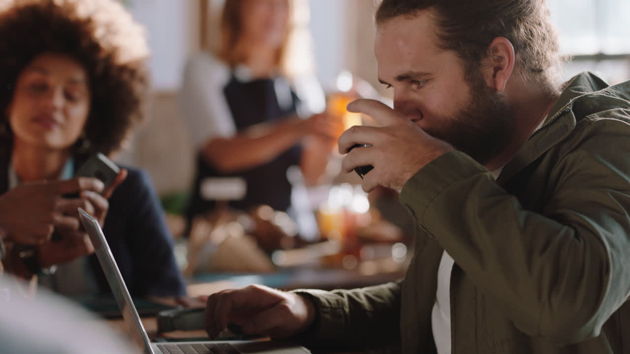 young caucasian man using laptop in cafe browsing online typing messages on smartphone drinking coffee in busy restaurant