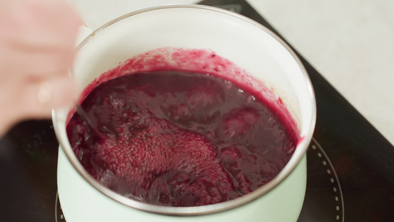 Close up of thick red soup or sauce bubbling in pastel green pot on electric stove while hand lifts twister utensil out, capturing texture, motion, and cooking heat in cozy home kitchen environment