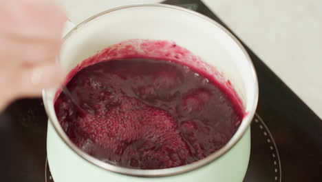 close up of thick red soup or sauce bubbling in pastel green pot on electric stove while hand lifts twister utensil out, capturing texture, motion, and cooking heat in cozy home kitchen environment