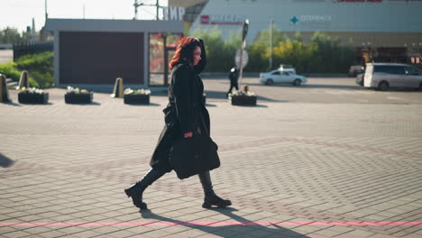 side view of businesswoman walking to office adjusting her hair while carrying black bag, dressed in black coat and boots, urban background features parked cars, office buildings, and pedestrians